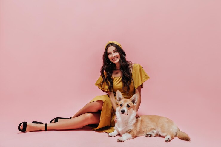 Foto grátis linda garota com cabelo comprido em vestido amarelo moderno e sapatos escuros sorrindo e posando com corgi em fundo rosa isolado