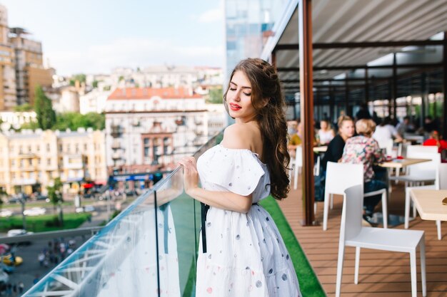 Linda garota com cabelo comprido fica no terraço no café. Ela usa um vestido branco com ombros nus e batom vermelho. Ela tem um sorriso leve e olha para baixo.