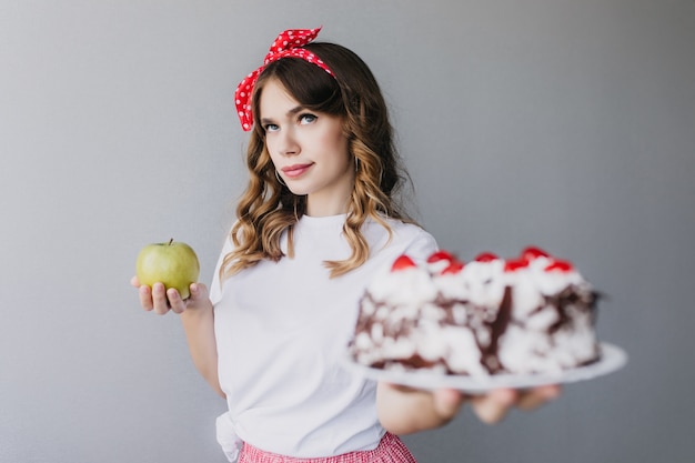 Linda garota com cabelo escuro ondulado, pensando em calorias e segurando o bolo. Foto interna de mulher jovem e bonita com torta de chocolate cremosa e maçã.