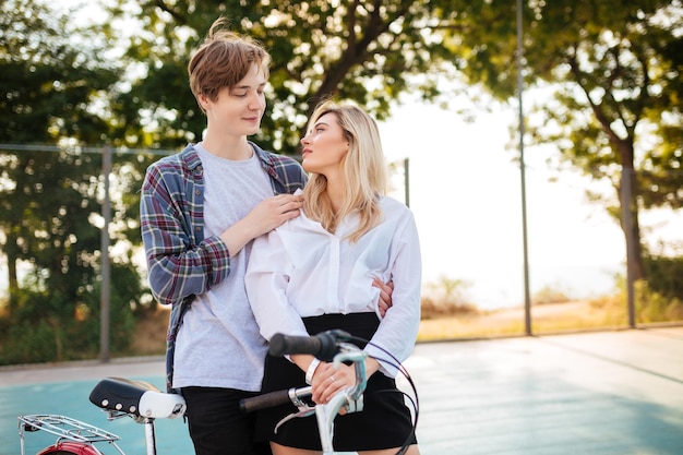 Linda garota com cabelo loiro e menino sonhando olhando um para o outro em pé no parque Retrato de um jovem casal fofo abraçando um ao outro com bicicleta ao lado