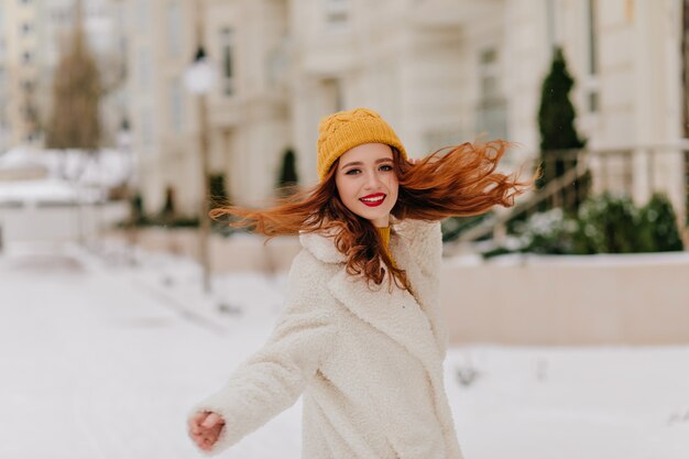 Linda garota com cabelo longo ondulado dançando na neve. Agradável modelo feminino com casaco se divertindo no inverno.