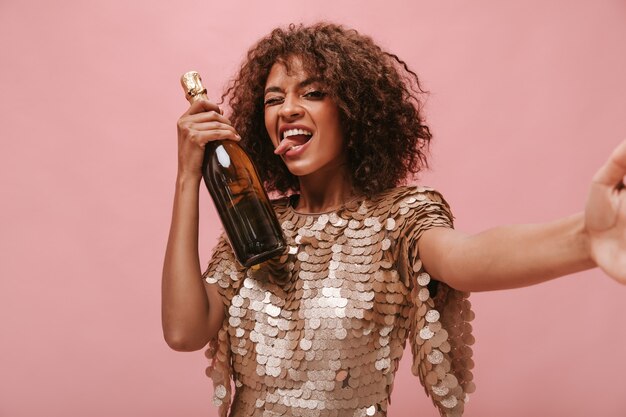 Linda garota com cabelo ondulado morena em um vestido brilhante piscando mostrando a língua, segurando a garrafa com a bebida e tirando foto na parede rosa.