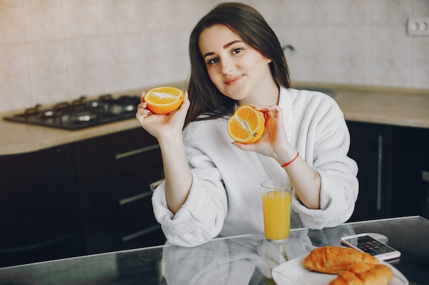 Linda garota com cabelo preto e roupão branco, sentado em casa na cozinha à mesa