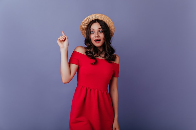 Linda garota com cabelos ondulados, posando na parede roxa com expressão de surpresa. Retrato interno de senhora bem-humorada em vestido vermelho e chapéu de palha.