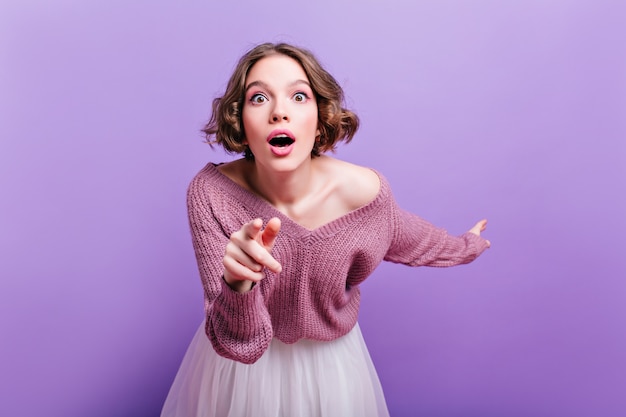 Linda garota com maquiagem rosa, expressando espanto e apontando o dedo. Foto interna de mulher chocada encaracolada isolada na parede roxa.