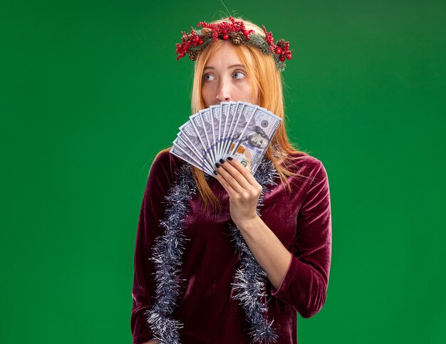 Linda garota com medo de olhar para o lado usando um vestido vermelho com coroa e guirlanda no pescoço coberto com dinheiro isolado no fundo verde