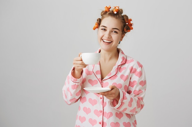 Linda garota com rolos de cabelo e pijama bebendo chá e sorrindo