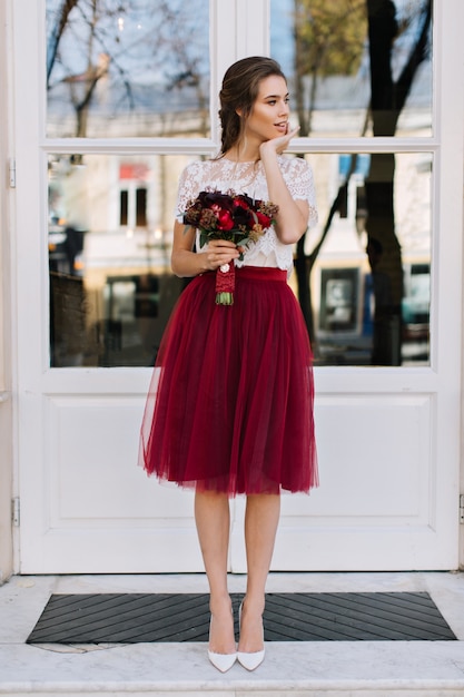 Foto grátis linda garota com saia de tule marsala nos saltos, andando na rua. ela segura um boquete de flores e sorri para o lado