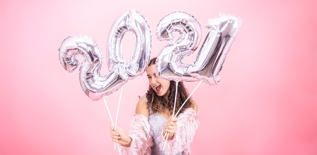 Linda garota com um sorriso em uma roupa festiva posando contra um fundo rosa do estúdio e segurando balões prateados para o conceito de ano novo