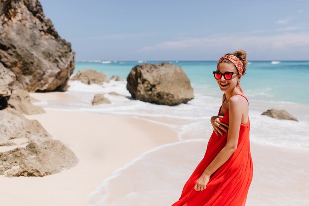 Linda garota com um sorriso encantador, passando a manhã perto do oceano. Foto ao ar livre de uma jovem europeia vestida de vermelho se divertindo no mar resort.