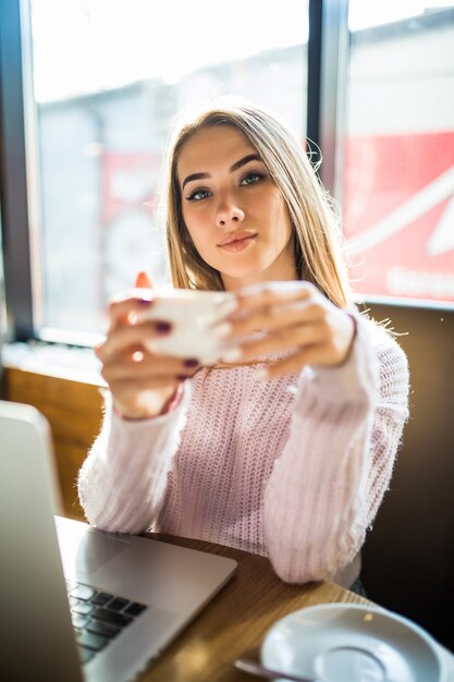 Linda garota com um suéter da moda, sentada em um café com uma xícara de chá, café, olhando para a câmera