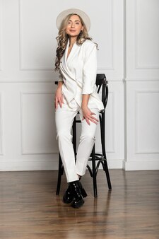 Linda garota com uma bela figura em um terninho branco e um chapéu branco de aba larga, posando contra um fundo branco no estúdio. sentado em uma cadeira