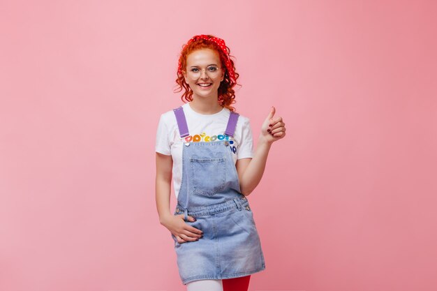 Linda garota com vestido de verão jeans e camiseta branca com um sorriso olhando para a câmera e mostrando os polegares para cima na parede isolada