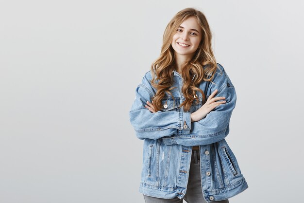Linda garota confiante em jaqueta jeans, sorrindo com os braços cruzados