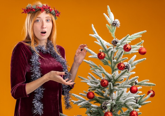 Linda garota confusa em pé perto da árvore de Natal, usando um vestido vermelho e uma grinalda com uma guirlanda no pescoço, isolada em um fundo laranja