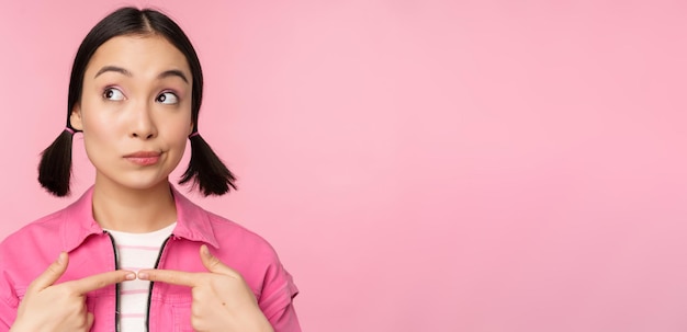 Foto grátis linda garota coreana com coques bobos parecendo feliz e tímido pensando em smth posando contra fundo rosa