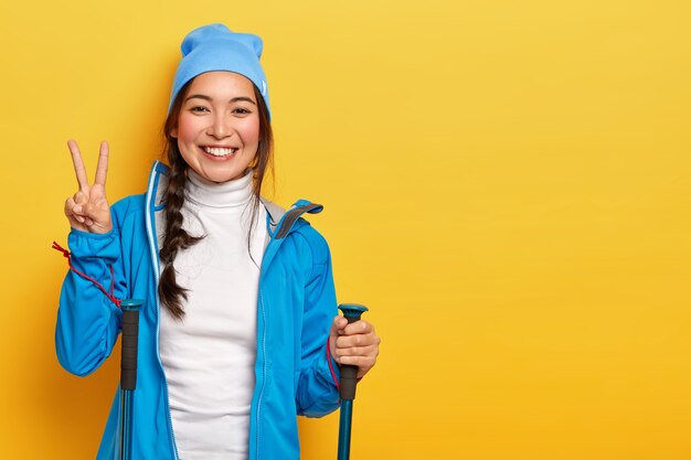 Linda garota coreana gosta de caminhadas, posa com bastões de trekking, faz gestos de paz, usa chapéu e jaqueta azul, isolado sobre fundo amarelo, espaço em branco