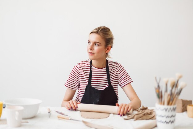 Linda garota de avental preto e camiseta listrada, sentado à mesa segurando o rolo trabalhando com argila e sonhadoramente olhando de lado no estúdio de cerâmica