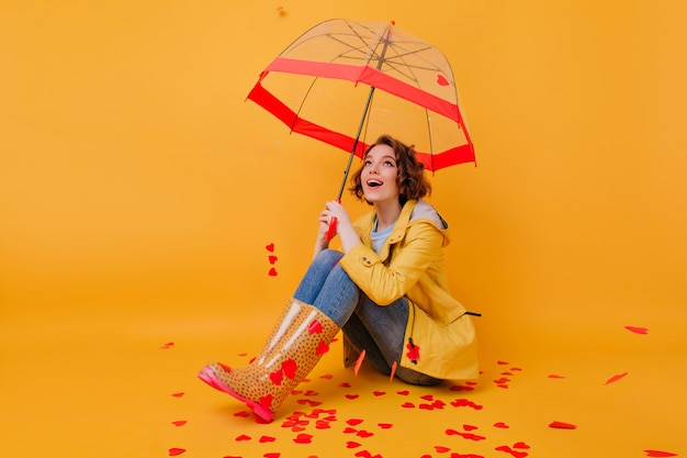 Linda garota de cabelo curto com belos olhos posando sob o guarda-sol. Foto interna do romântico modelo feminino branco sentado no chão amarelo com guarda-chuva.