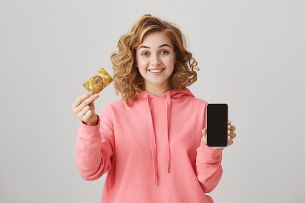 Linda garota de cabelos cacheados mostrando um cartão de crédito dourado e a tela do celular
