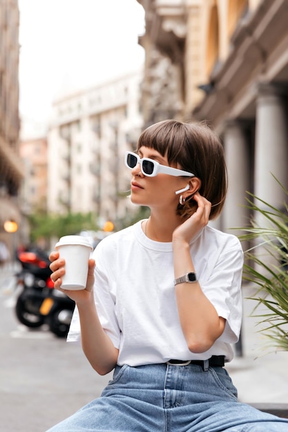 Linda garota de cabelos escuros em óculos de sol bebendo café em bom dia ensolarado Retrato de modelo feminino jocund em roupa elegante segurando café