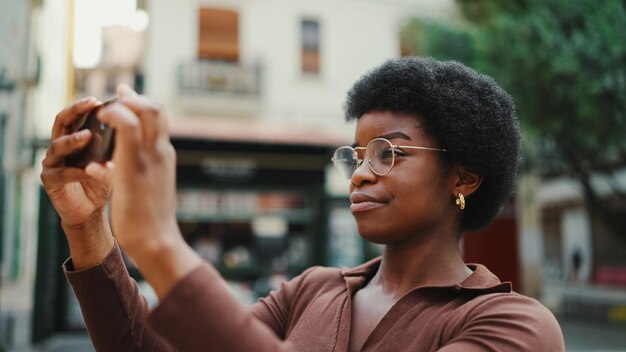 Linda garota de cabelos escuros em óculos tirando foto para seu blog ao ar livre mulher afro usando smartphone na rua