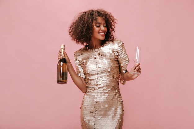 Linda garota de cabelos fofos em um elegante vestido brilhante, sorrindo com os olhos fechados e segurando o copo e a garrafa com vinho na parede isolada.