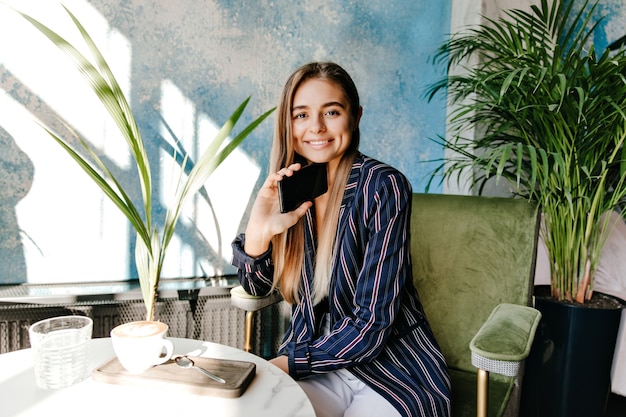 Linda garota de casaco azul, aproveitando a pausa para o café. Mulher de cabelos compridos rindo, sorrindo em um café aconchegante.