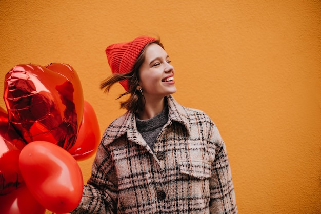 Linda garota de chapéu elegante está sorrindo sinceramente contra a parede laranja Mulher se alegra no dia dos namorados e posa com balões em forma de coração