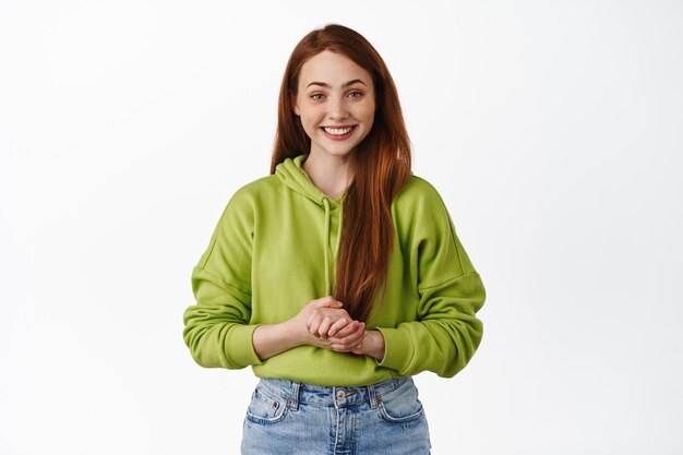 Linda garota de faculdade ruiva, parece esperançosa e sorrindo para a câmera, de mãos dadas juntas de maneira educada e amigável, ajudando a ajudar, de pé sobre fundo branco.