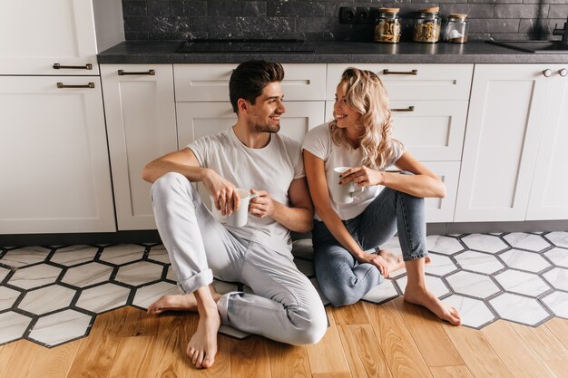 Linda garota de jeans, sentada no chão e conversando com o namorado. Jovem casal desfrutando de café na cozinha.