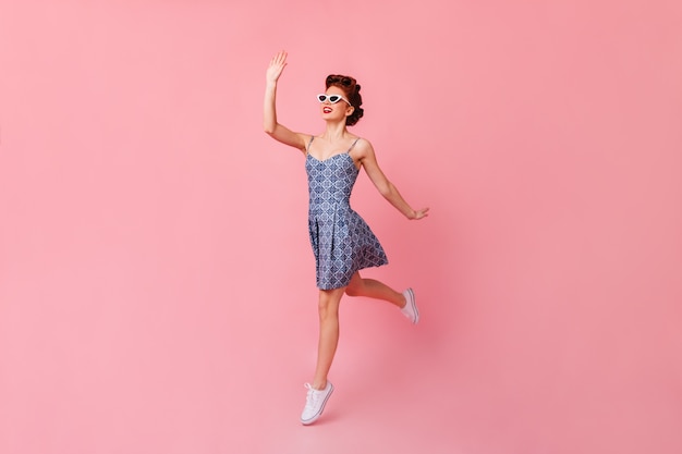 Linda garota de óculos de sol acenando com a mão. Foto de estúdio de feliz mulher pinup pulando no espaço rosa.