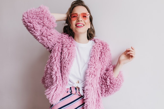 Linda garota de óculos de sol rosa posando com um sorriso alegre na parede branca. Retrato interior de mulher de cabelo curto com casaco de pele, brincando com seu cabelo.