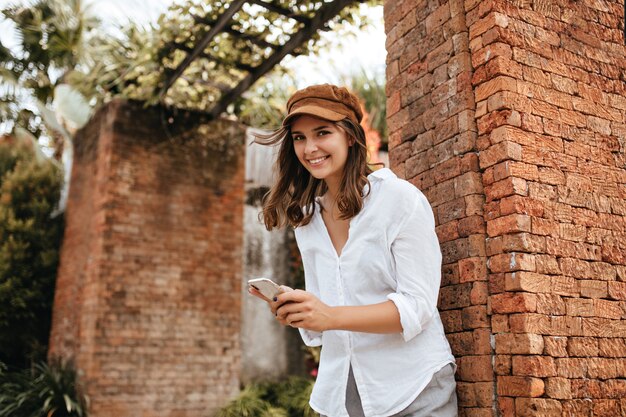 Linda garota de olhos castanhos com sorriso posa ao lado do prédio de tijolos. Mulher de boné e camisa branca, segurando o smartphone.