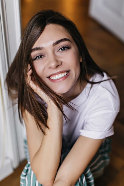 Linda garota de olhos escuros sorrindo enquanto relaxa em casa. Foto aérea interna de mulher atraente morena em camiseta branca.