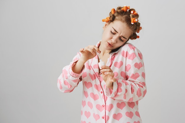 Foto grátis linda garota de pijama e bobes de cabelo, falando ao telefone enquanto aplica esmalte