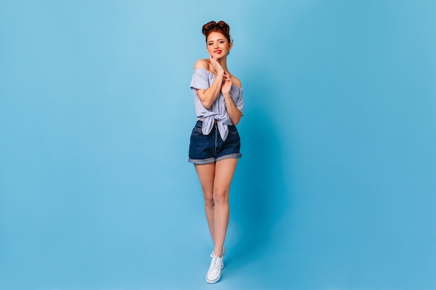 Linda garota de shorts jeans e blusa de pé no espaço azul. visão de comprimento total da senhora pin-up bem torneada.