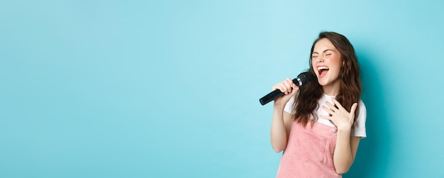 Linda garota despreocupada cantando no microfone com paixão tocando karaokê em pé sobre fundo azul