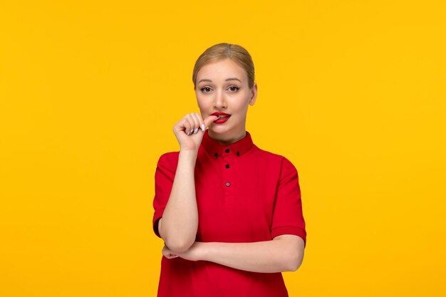 Linda garota do dia da camisa vermelha mordendo a unha em uma camisa vermelha e batom em um fundo amarelo