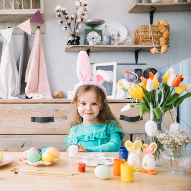 Linda garota em orelhas de coelho sentado à mesa com ovos coloridos
