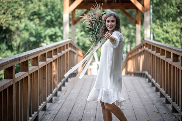 Linda garota em um vestido branco com um buquê de flores exóticas em uma ponte de madeira.