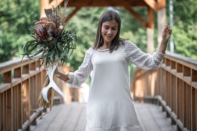 Linda garota em um vestido branco com um buquê de flores exóticas em uma ponte de madeira.