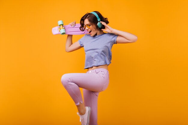 Linda garota encaracolada em calças rosa pulando enquanto ouve música. Foto interna de feliz senhora caucasiana em fones de ouvido, dançando com o skate.