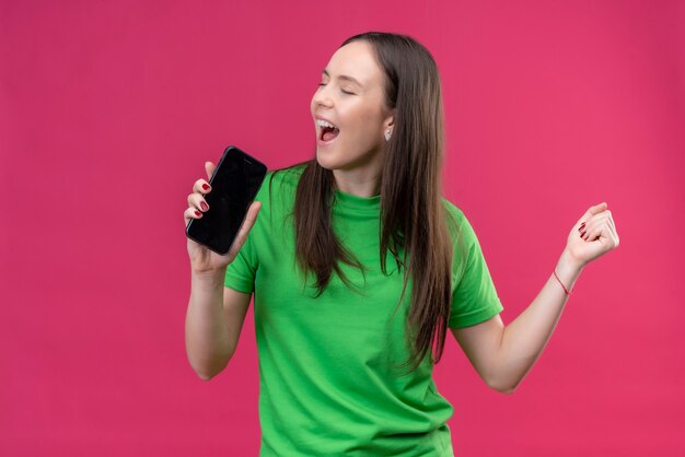 Linda garota engraçada vestindo uma camiseta verde segurando um smartphone e usando-o como microfone cantando em pé sobre o espaço rosa isolado