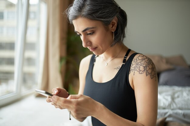 Linda garota esportiva com cabelo grisalho prematuro, tatuagem no ombro e piercing no nariz, sentada dentro de casa com um telefone celular