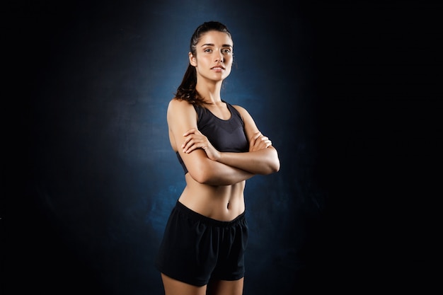 Foto grátis linda garota esportiva posando com os braços cruzados sobre parede escura.