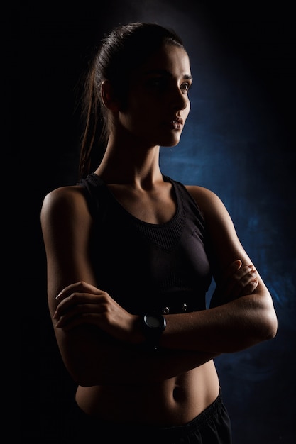 Linda garota esportiva posando com os braços cruzados sobre parede escura.