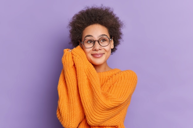 Linda garota étnica com cabelo afro natural mantém as mãos perto do rosto e parece com uma expressão sonhadora em algum lugar usa um suéter de malha laranja solto.