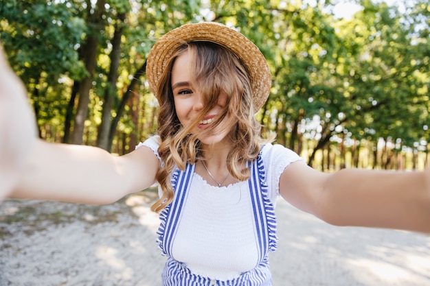 Linda garota europeia com cabelos ondulados posando de brincadeira no parque. senhora encantadora de chapéu fazendo selfie com árvores
