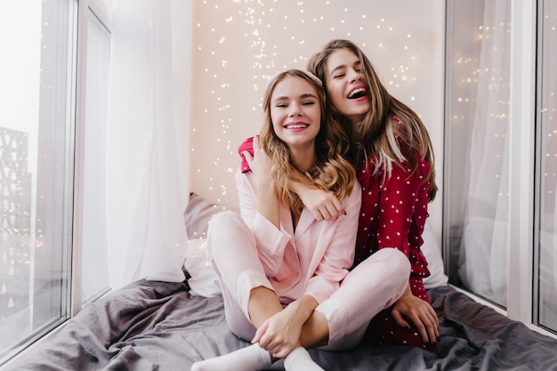 Foto grátis linda garota europeia em pijamas rosa, posando com um sorriso encantador no quarto. retrato interno de mulheres fascinantes de pijama se abraçando e rindo.